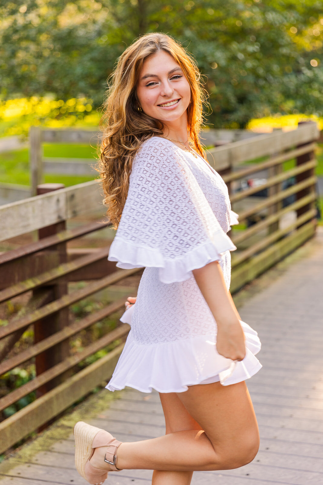 senior girl in short white dress looking at the camera over her shoulder one leg up on a Sandy Spring park bridge during photo session