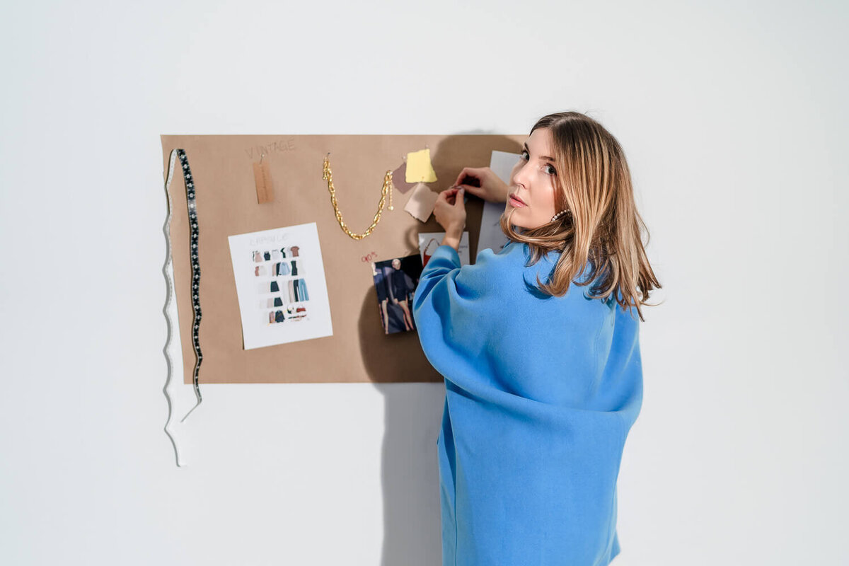 Woman in blue sweater arranging fabric swatches on inspiration board with gold necklace and photos.