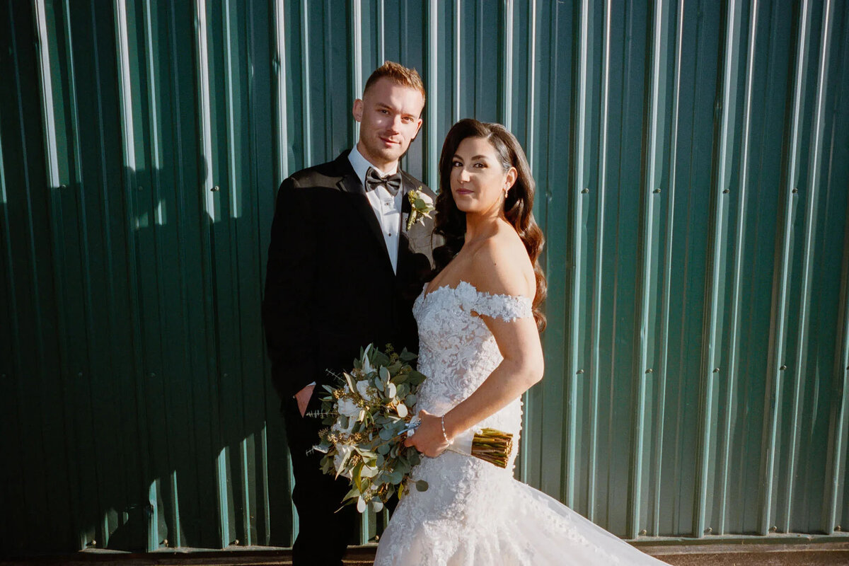 A bride in an off-shoulder white gown holds a bouquet and stands beside a groom in a black tuxedo. Captured by a film photographer NJ, they pose together before a green corrugated metal wall, facing the camera.