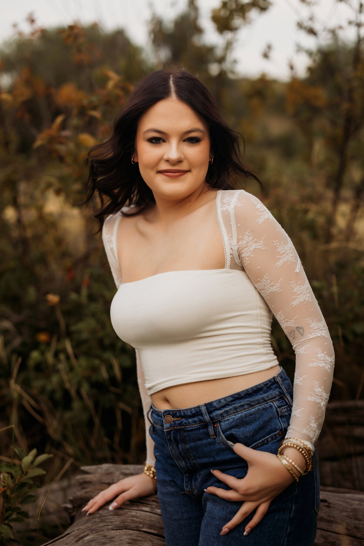 HIgh school senior girl poses against a fallen tree stump with colorful leaves behind her for her senior photo session