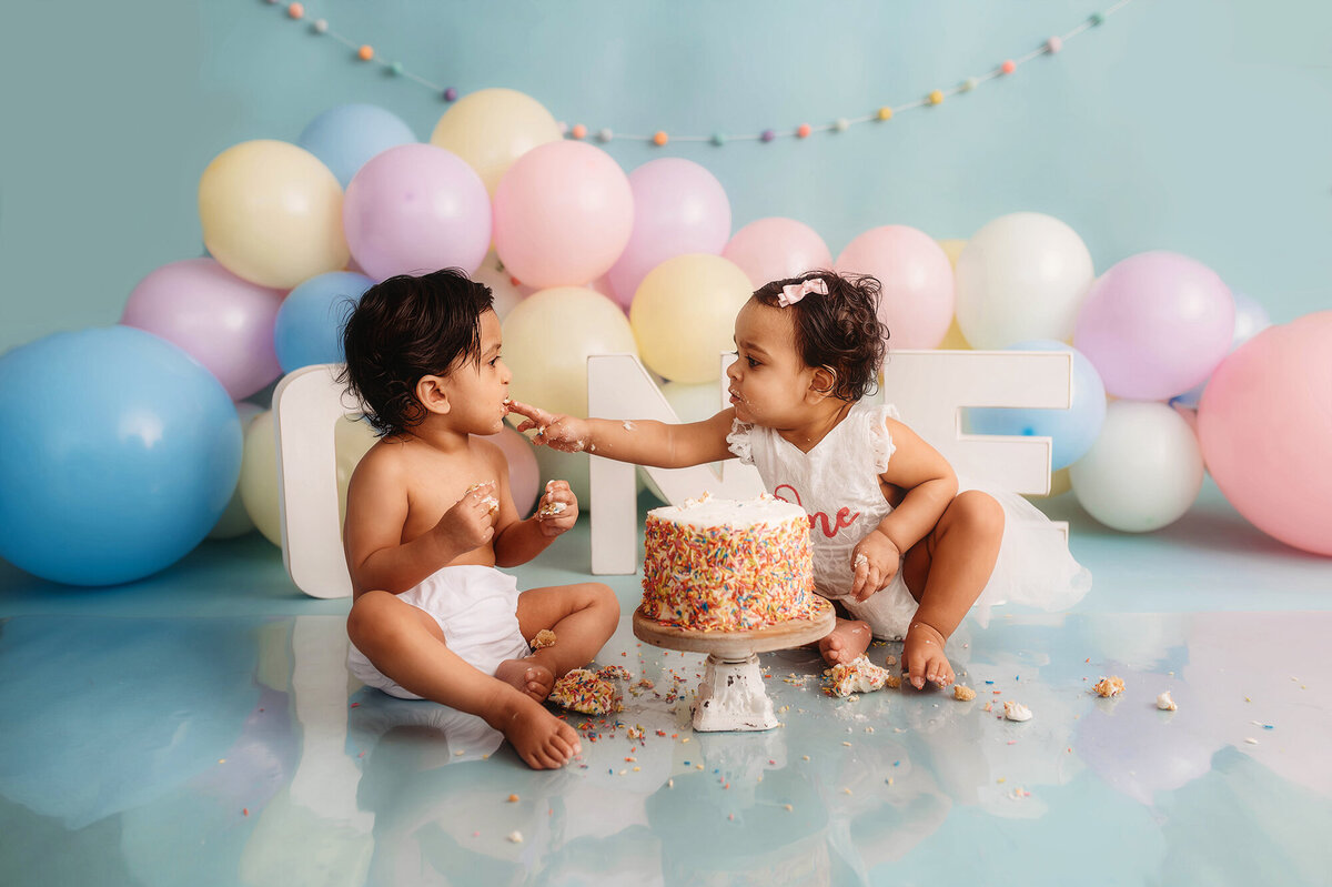 Twin babies feed each other birthday cake during cake smash portrait session in Asheville, NC photo studio.