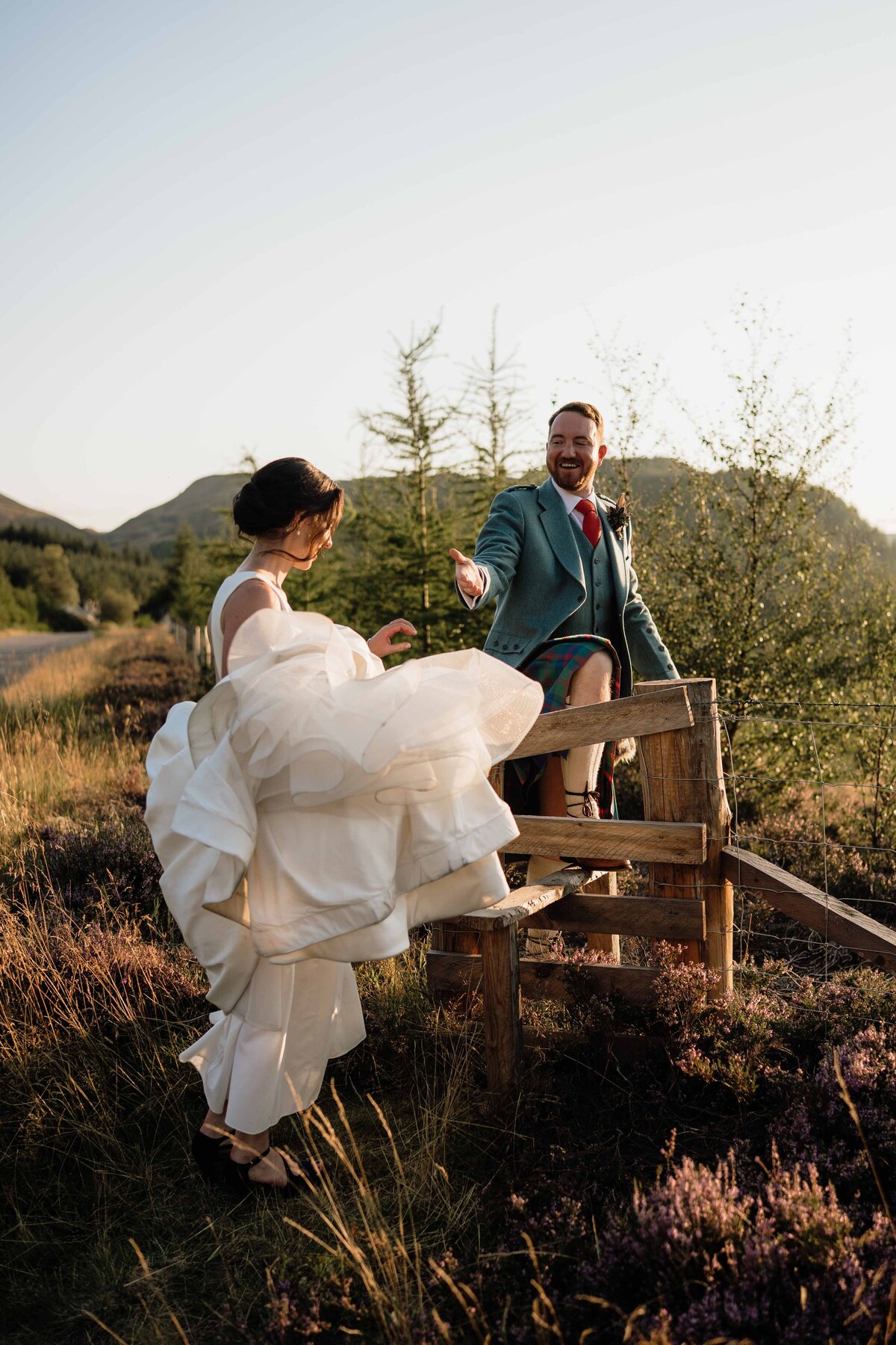 A couple are photographed waring wedding attire and climbing over a wooden stile.