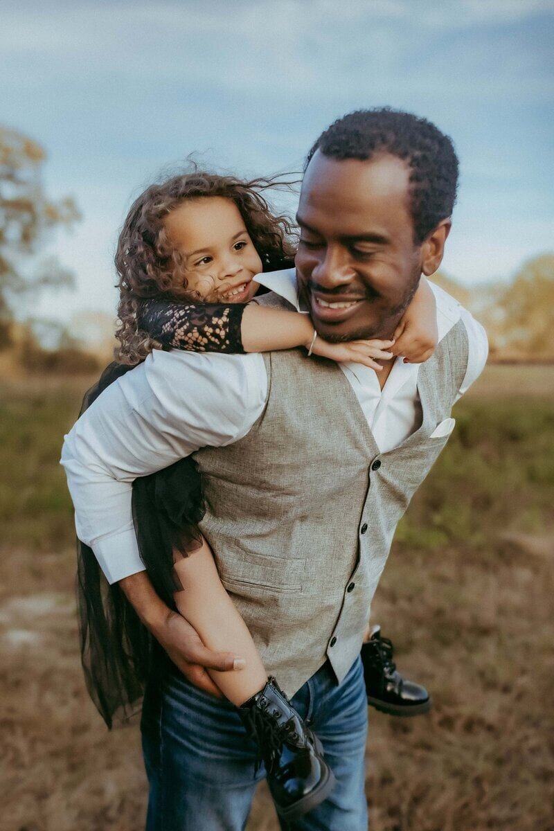 A man wearing a vest is giving a young girl a piggyback ride in an open field. Both are smiling, and the girl is wearing a black dress and boots. The sky is clear.