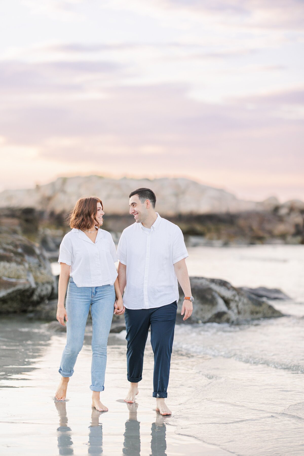 A man and woman holding hands walking down Wingaersheek Beach during their sunset engagement portraits.