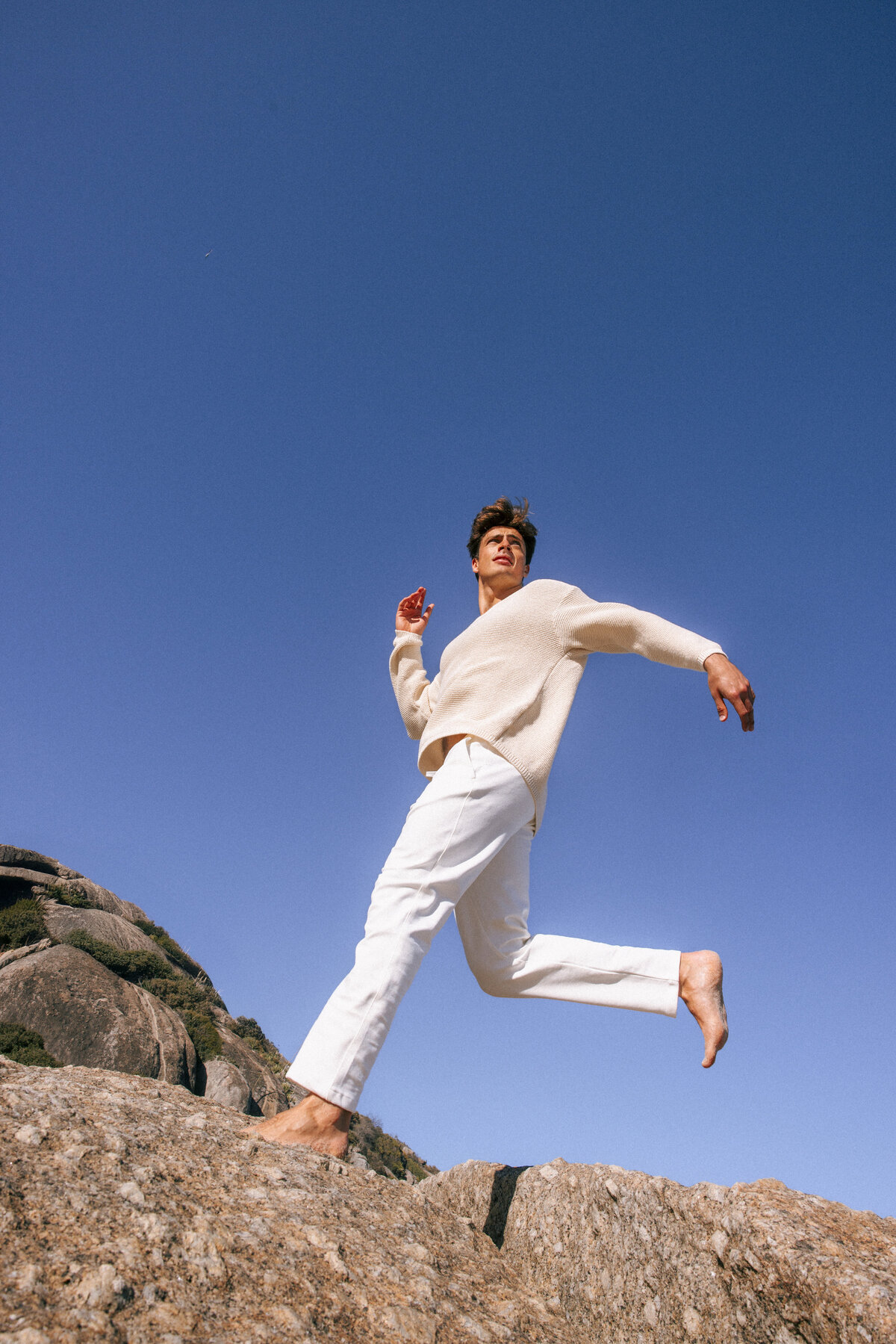 Male model in a cream sweater and white trousers mid-leap on a rocky surface in Cape Town, South Africa. Captured against a vibrant blue sky, evoking energy and freedom in a coastal fashion editorial.