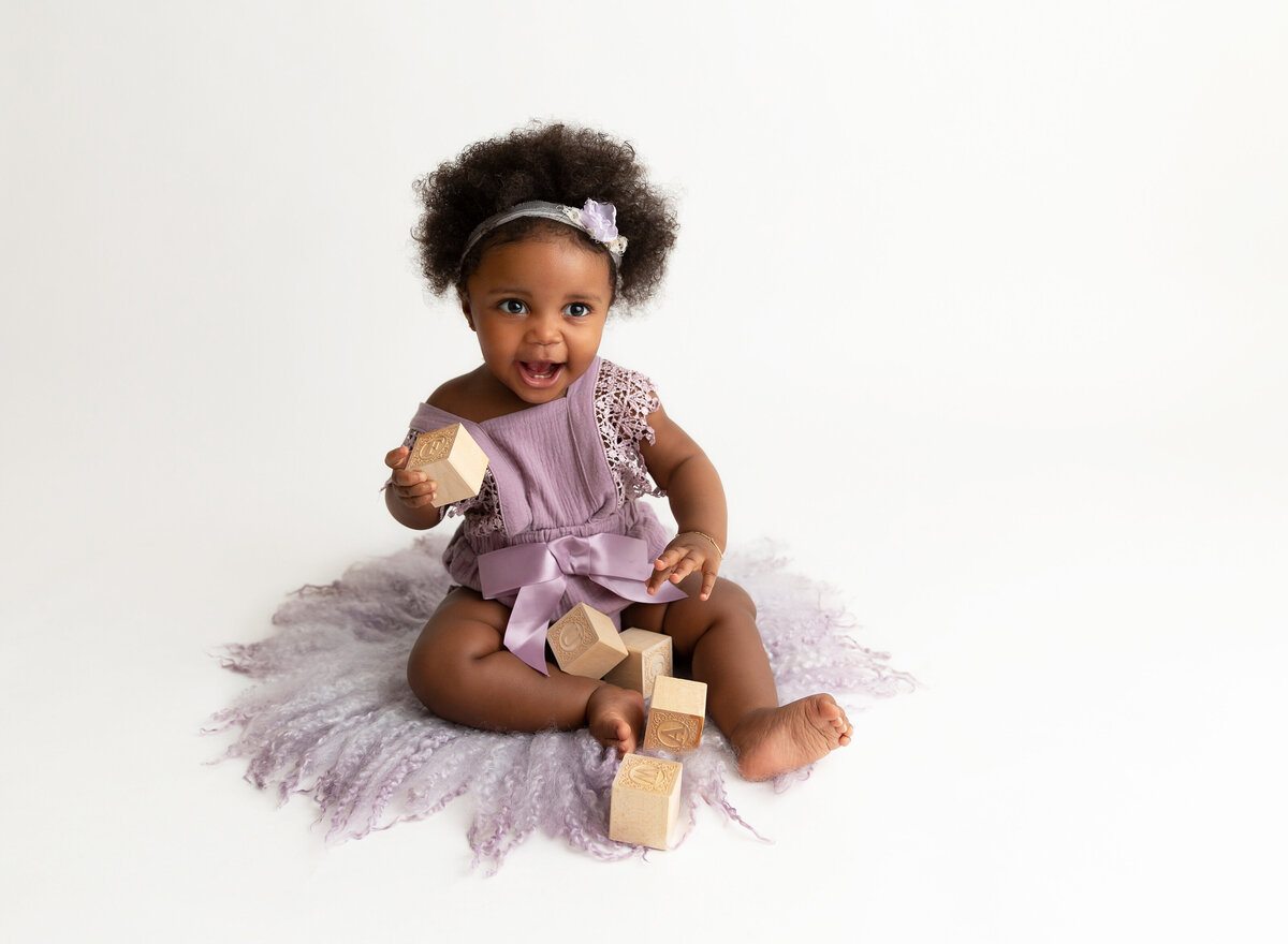 Baby girl sitting for a 6-month baby milestone photo ssession. Baby girl is in a purple knit romper and playing with stacked wooden blocks. Baby is smiling at the camera.