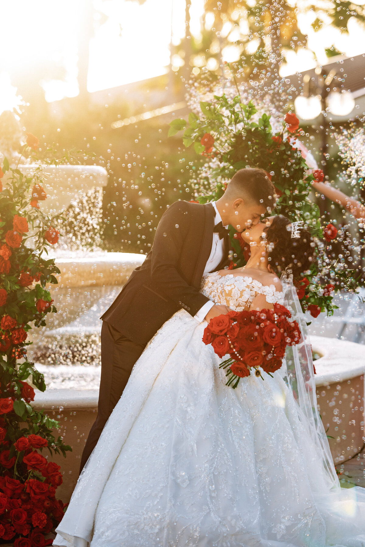 Shannon Lee, from Love Like Wild Photography, captures a couple sharing a romantic kiss by a fountain, surrounded by red roses.