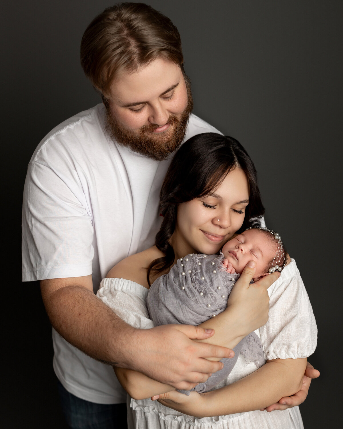 Mom and dad embracing their newborn daughter, wrapped in a gray knit outfit with delicate pearl accents, during their studio session. Captured by Inland Empire newborn photographer Kaitlyn Dawn Photography.