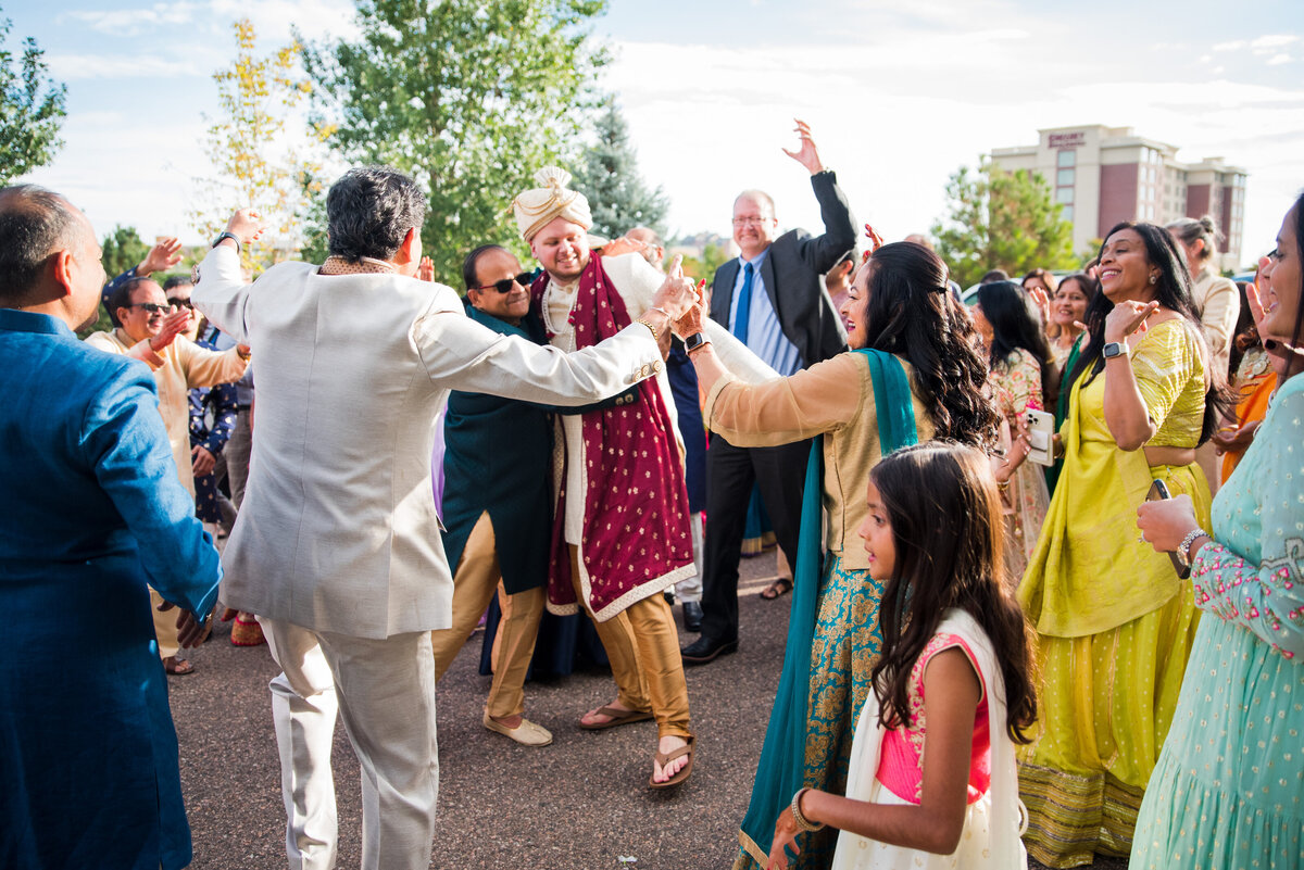 A groom in a traditional Indian outfit is in a crowd of family and friends being celebrated.