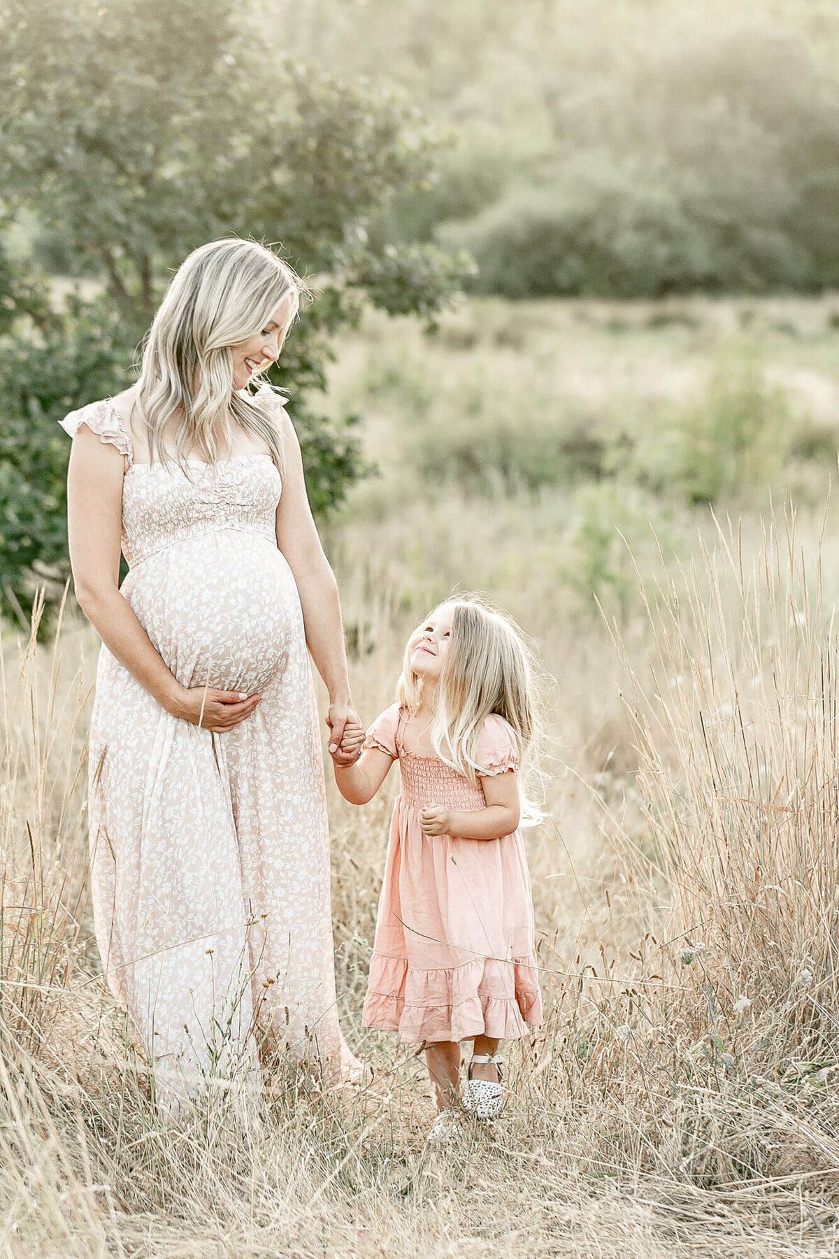 Mom holding her baby bump with one hand and holding hands with her toddler girl. She and her toddler are looking  at each other and walking in a field of tall golden grasses.