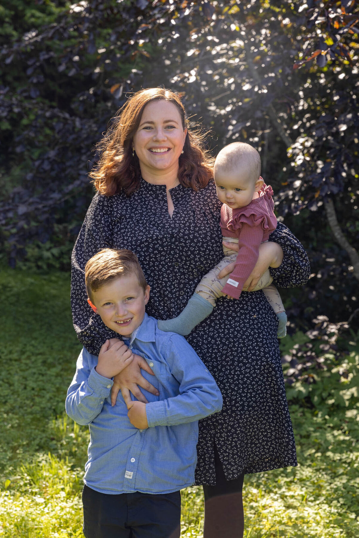 Familiefoto av mor og to barn; eldstegutten står foran henne og holder rundt mors arm, barn 2 er på mors hofte. Fotografert av Linn Landmark.