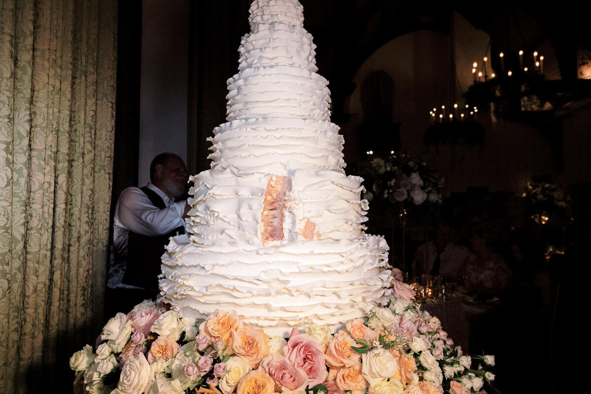 A huge, white, six-layer wedding cake with many flowers at the bottom, and a slice in the middle of the second layer.
