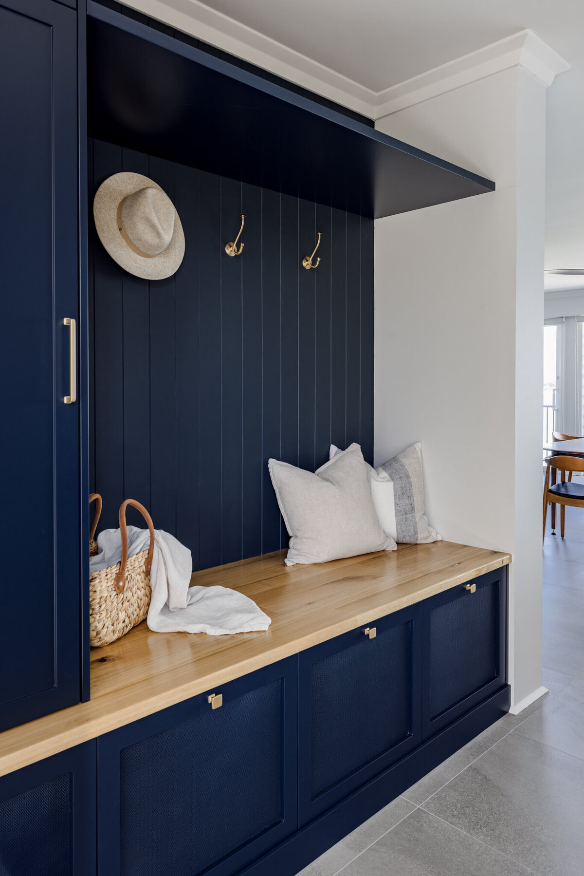 A mudroom nook with a wooden bench, navy cabinets, hooks, a hat, a wicker basket, and white pillows showcases the elegant touch of Newcastle Interior Designer expertise.