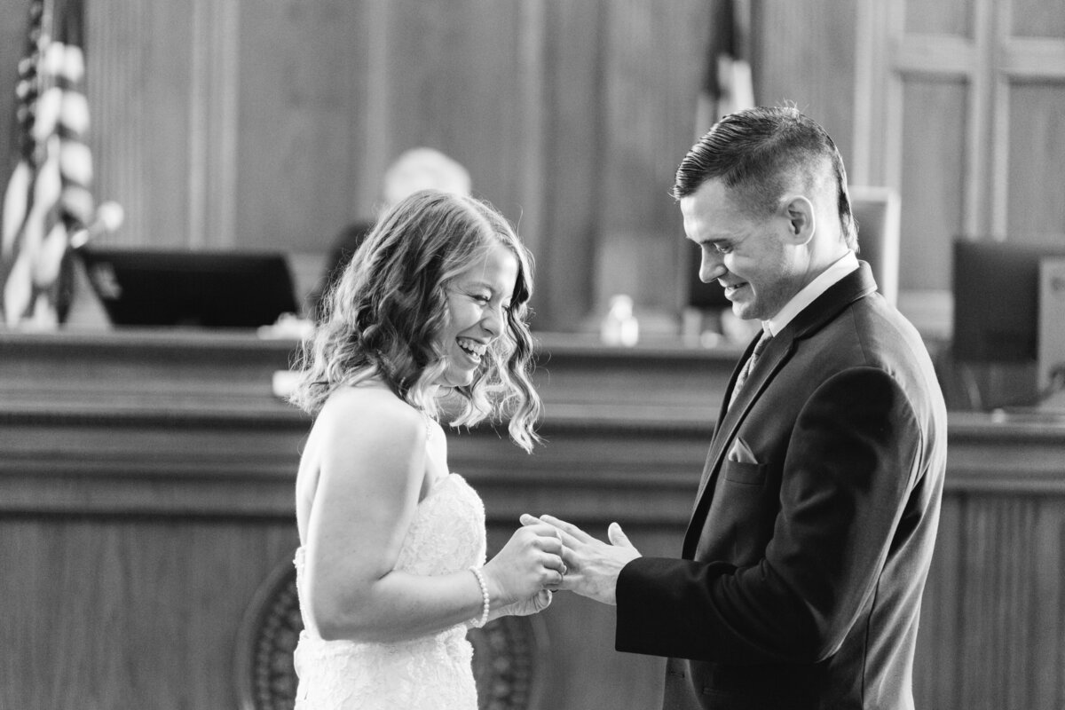 Black and white candid shot of a bride joyfully putting a wedding ring on her groom's ring finger during their wedding ceremony at the Tarrant County Courthouse in Fort Worth, Texas. The bride is wearing a sleeveless, white dress while the groom is wearing a suit. A judge can partially be seen in the background.