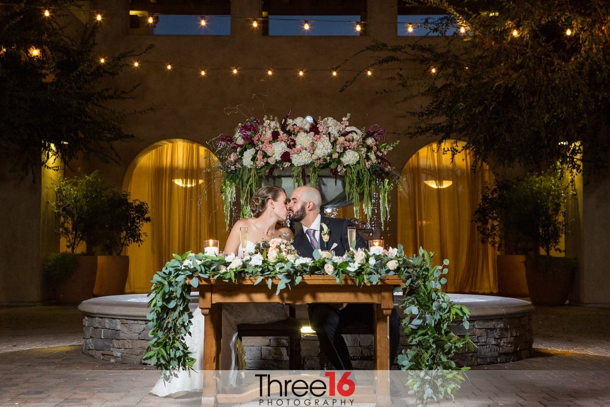 Bride and Groom share a kiss while sitting at the floral sweetheart table prior to dinner