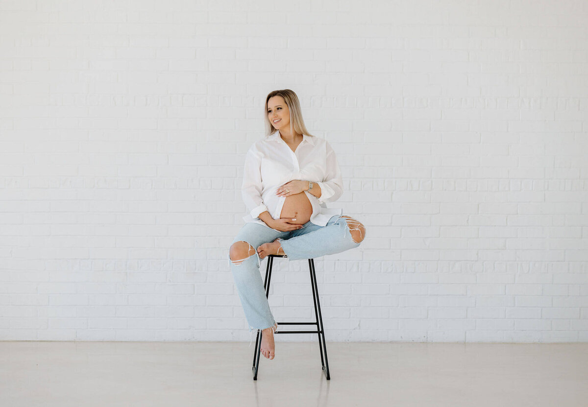 fine art maternity portrait of expecting mom in white button up shirt and jeans sitting on stool in Tyler Texas Studio