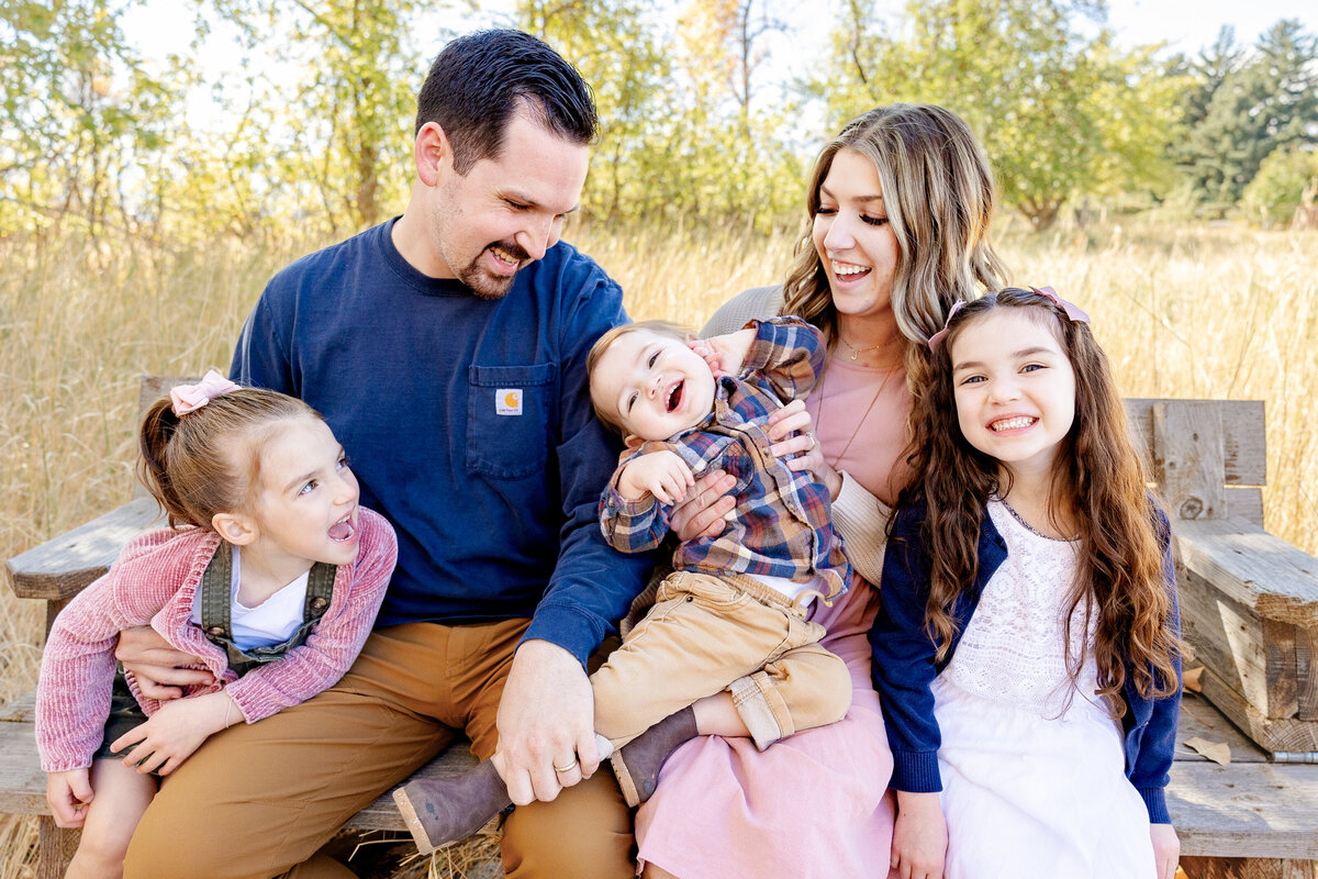 A family of five sits on a rustic wooden bench, playfully tickling their youngest child. The warm autumn light and golden grasses frame this heartwarming moment of love and laughter.
