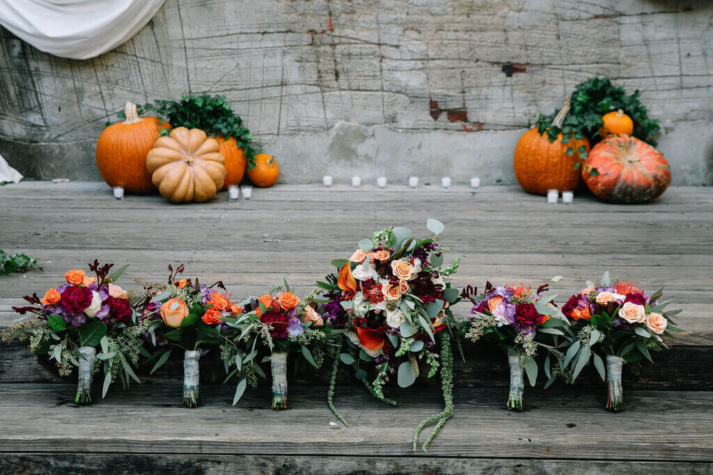 Several vibrant floral bouquets featuring roses, eucalyptus, and other colorful flowers are lined up on wood stairs. Pumpkins and candles decorate the background.