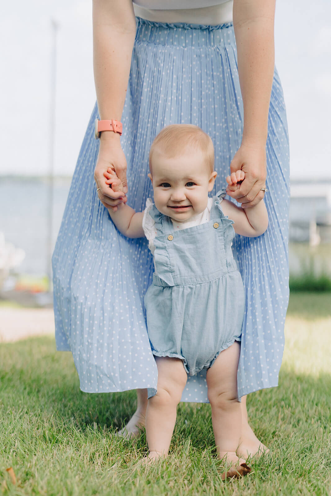 Baby walking with arms held up by woman in blue skirt