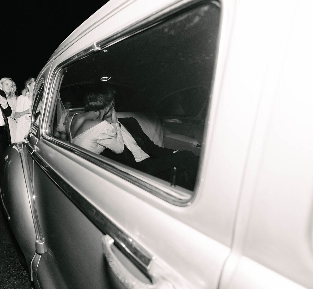 Bride and groom embrace and kiss inside their vintage getaway car, photographed through the window during their wedding exit in Highlands, NC.