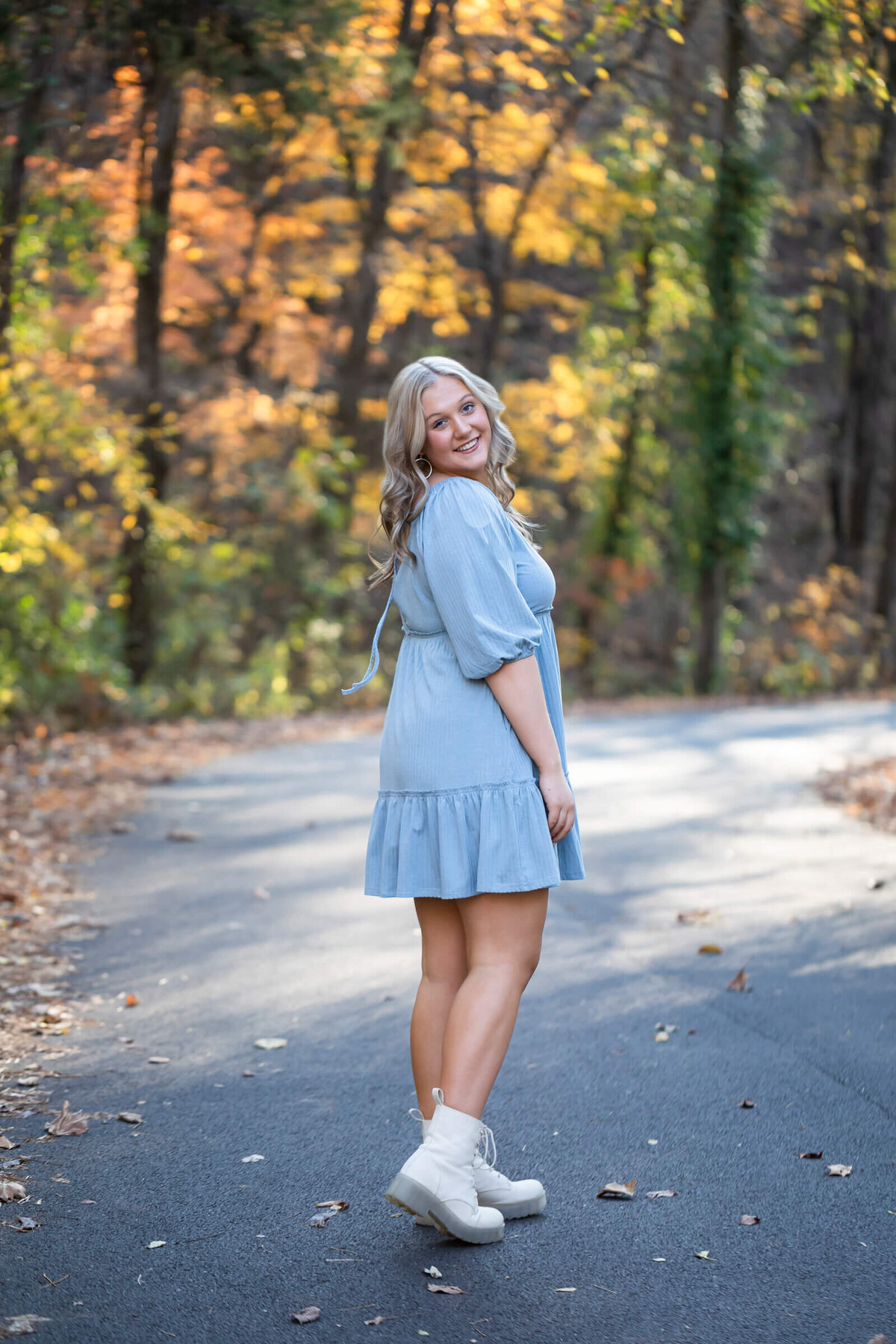 A pretty blonde haired teen girl in a baby blue dress spins on a country road surrounded by colorful fall leaves. Photograph by Dynae Levingston.