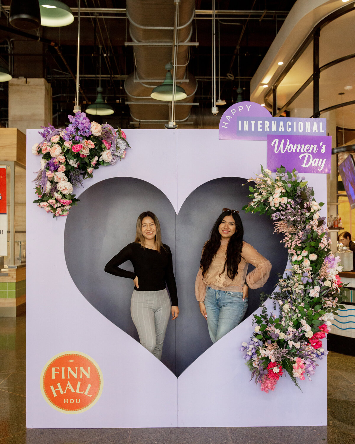 Two women posing in a purple heart shaped photo booth backdrop display decorated with florals for International Women's Day