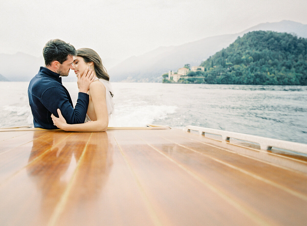 Film photograph of bride and groom on a wooden boat on Lake Como in Italy she is wearing a white halter jumpsuit with her hair down in loose waves and he is wearing grey and navy cashmere sweater and coat photographed by Italy wedding photographer