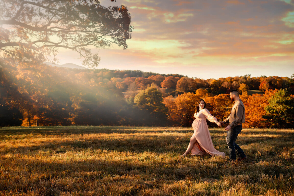 An expecting couple holds hands and walks across a meadow during sunset by the Biltmore Estate