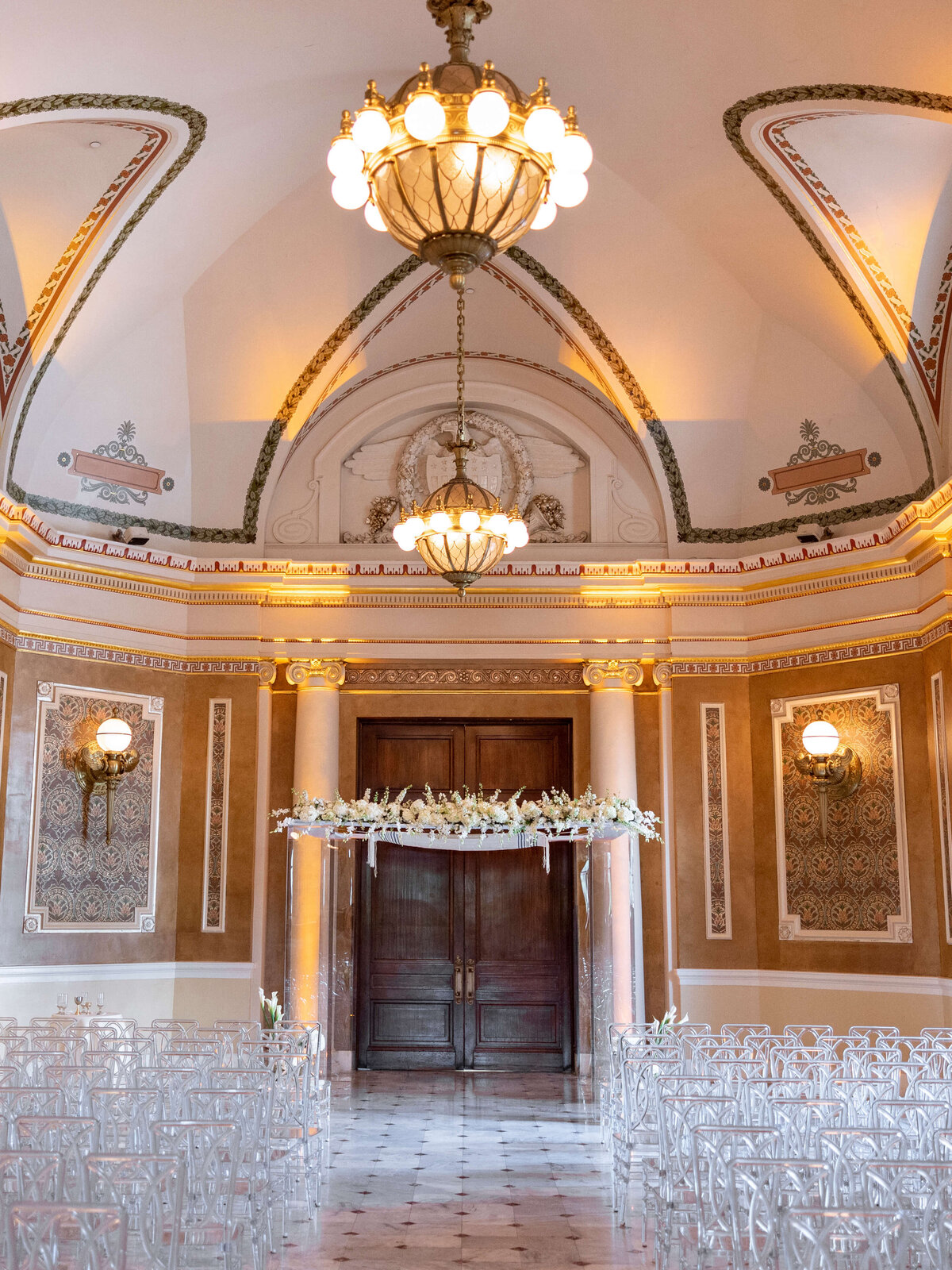 Ornate wedding venue with high arched ceilings, chandeliers, and intricate wall designs. Clear chairs face a dark wooden door framed by floral decorations. Marble floors add elegance to the setting.