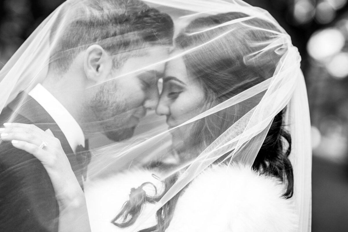 A black-and-white image of the groom and bride sharing a tender moment under the bride's veil. The groom smiles lovingly at the bride, who is also smiling. The soft light and intimate setting highlight their connection, with the veil adding a romantic and ethereal touch to the image.