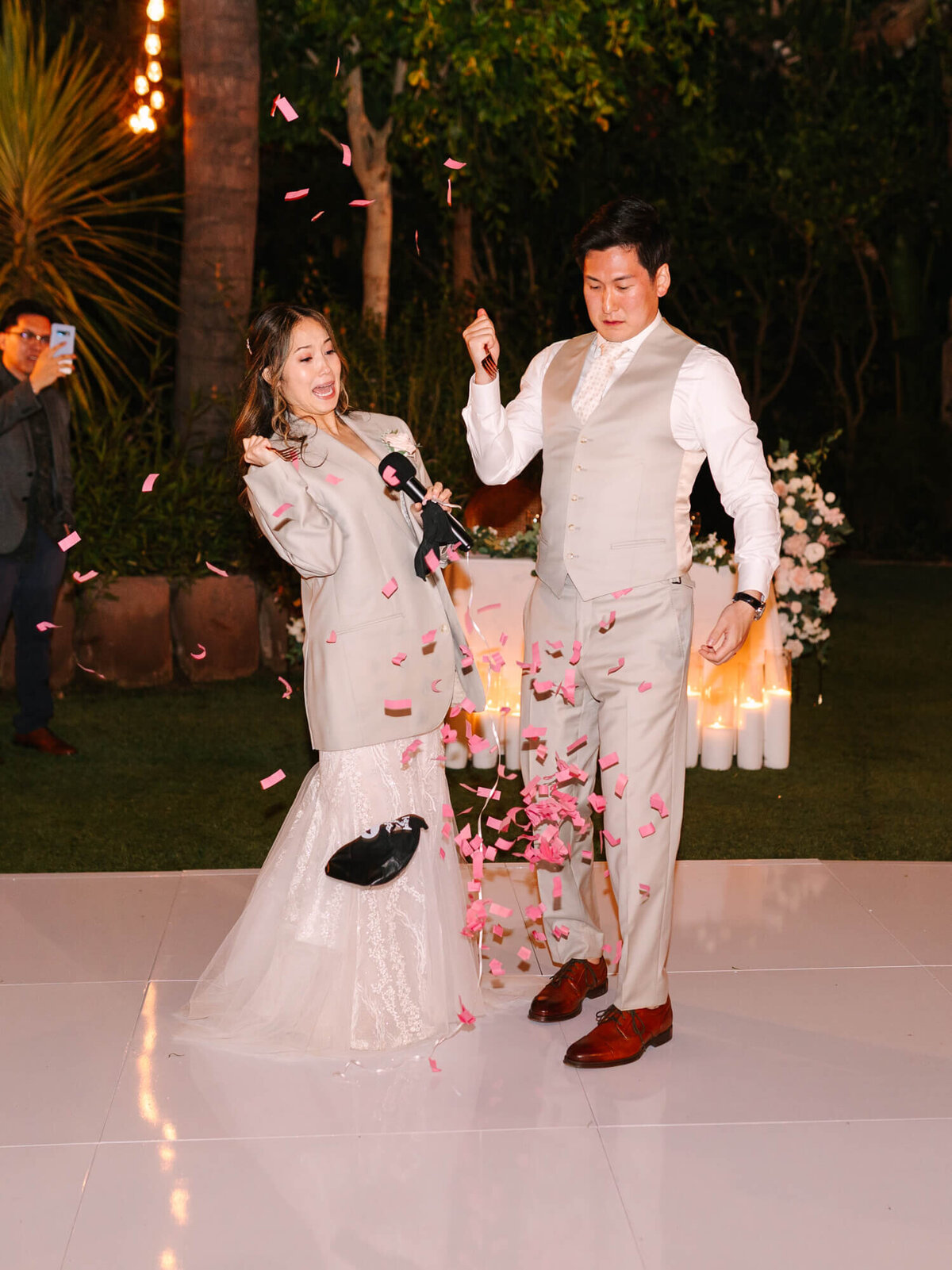 A joyful bride and groom in suits pop open a balloon on a lit floor, surrounded by falling pink confetti.