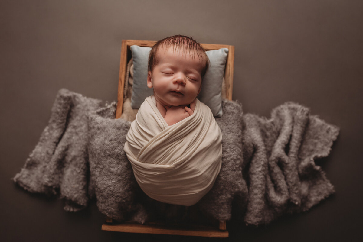 Baby boy 2 weeks old asleep posed by Woodstock GA newborn photographer in wooden drawer wrapped in cream swaddle and laying on gray cloth