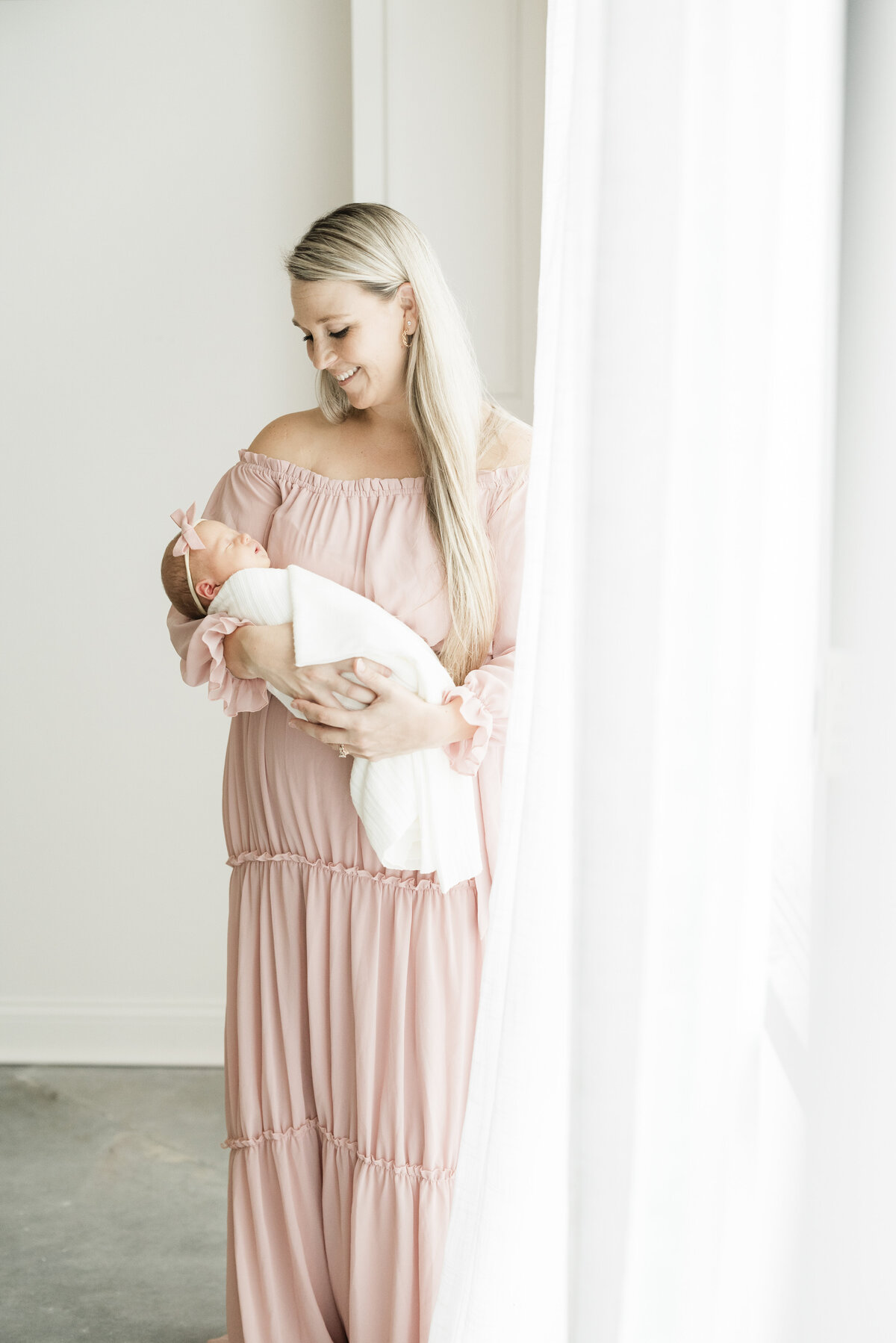 Mother standing near sheer white curtains while gently holding her newborn during a natural light Kansas City newborn session.