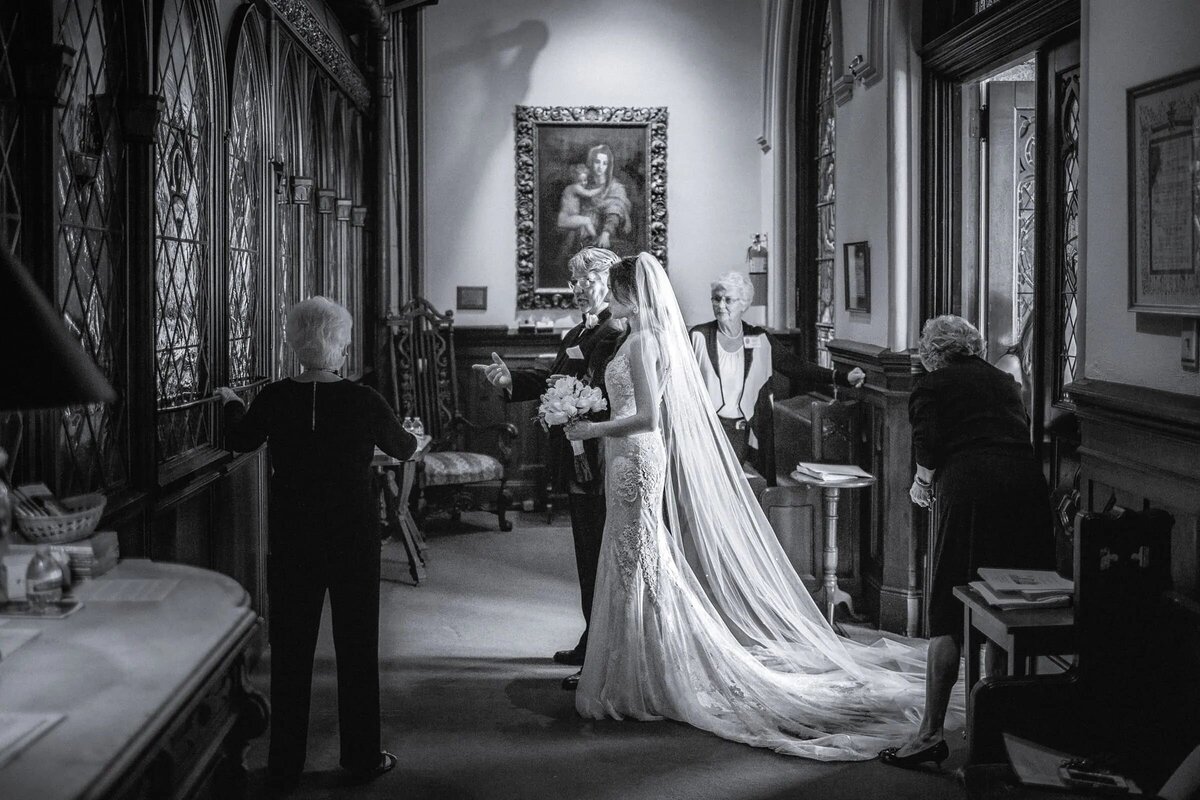 Elegant black and white photo of a bride making an entrance, her train and veil beautifully highlighted, with older individuals assisting