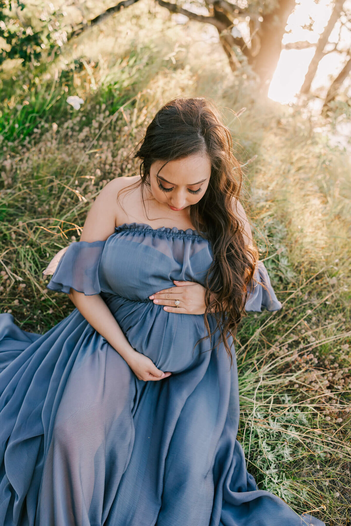 portrait of a woman sitting on the hill in a blue dress with her hands on her belly. She is looking down at her belly and her dress is flowing off to the side.