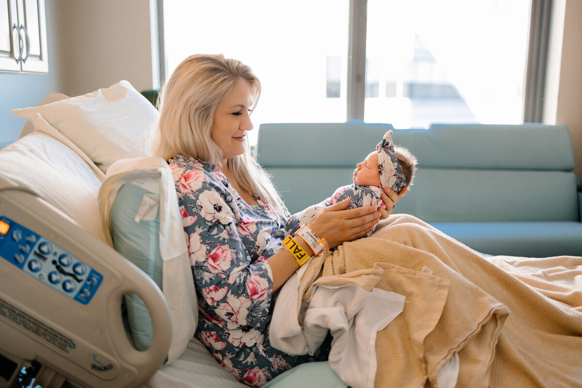 Mother holding her newborn baby at the hospital, sitting on a couch with pillows. She is wearing a blue floral shirt and a light brown blanket. The baby, with a blue floral headband, is peacefully sleeping.