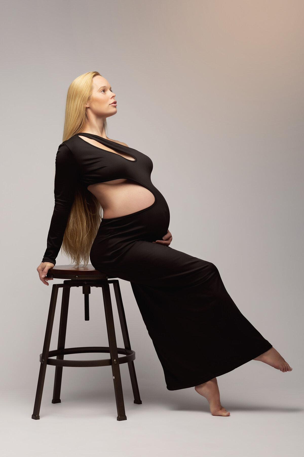 A striking portrait of an expecting mother dressed in a sleek, black cut-out gown, elegantly seated on a stool with her body slightly arched. Her long blonde hair flows over her shoulder, and her calm expression conveys the strength and beauty captured by DFW maternity photography expert Lindsay Walden.