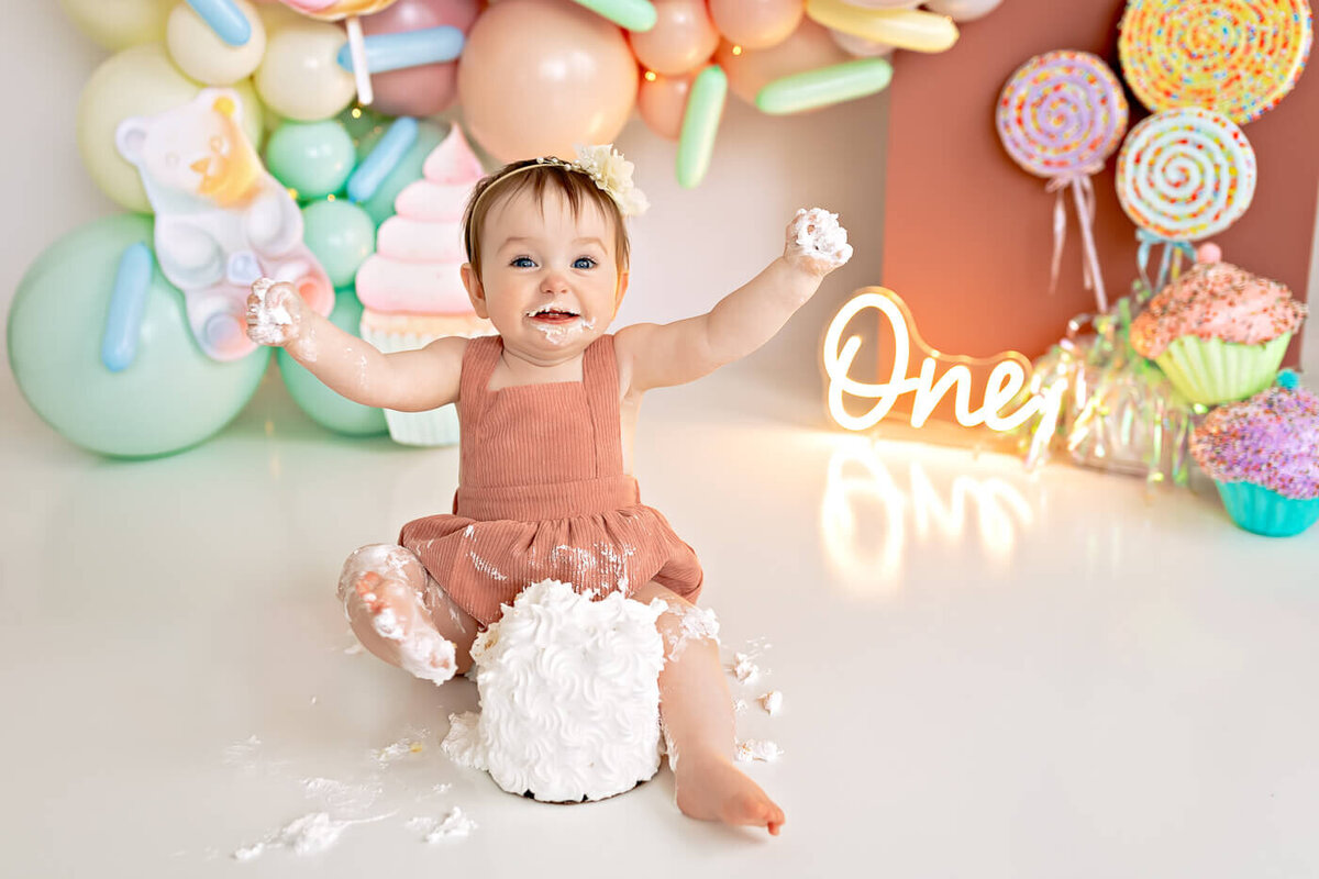 A baby sits joyfully on the floor covered in cake frosting, holding some in each hand. Behind, colorful balloons, candies, and a neon "One" sign celebrate a first birthday. The baby wears a brown outfit and a headband.