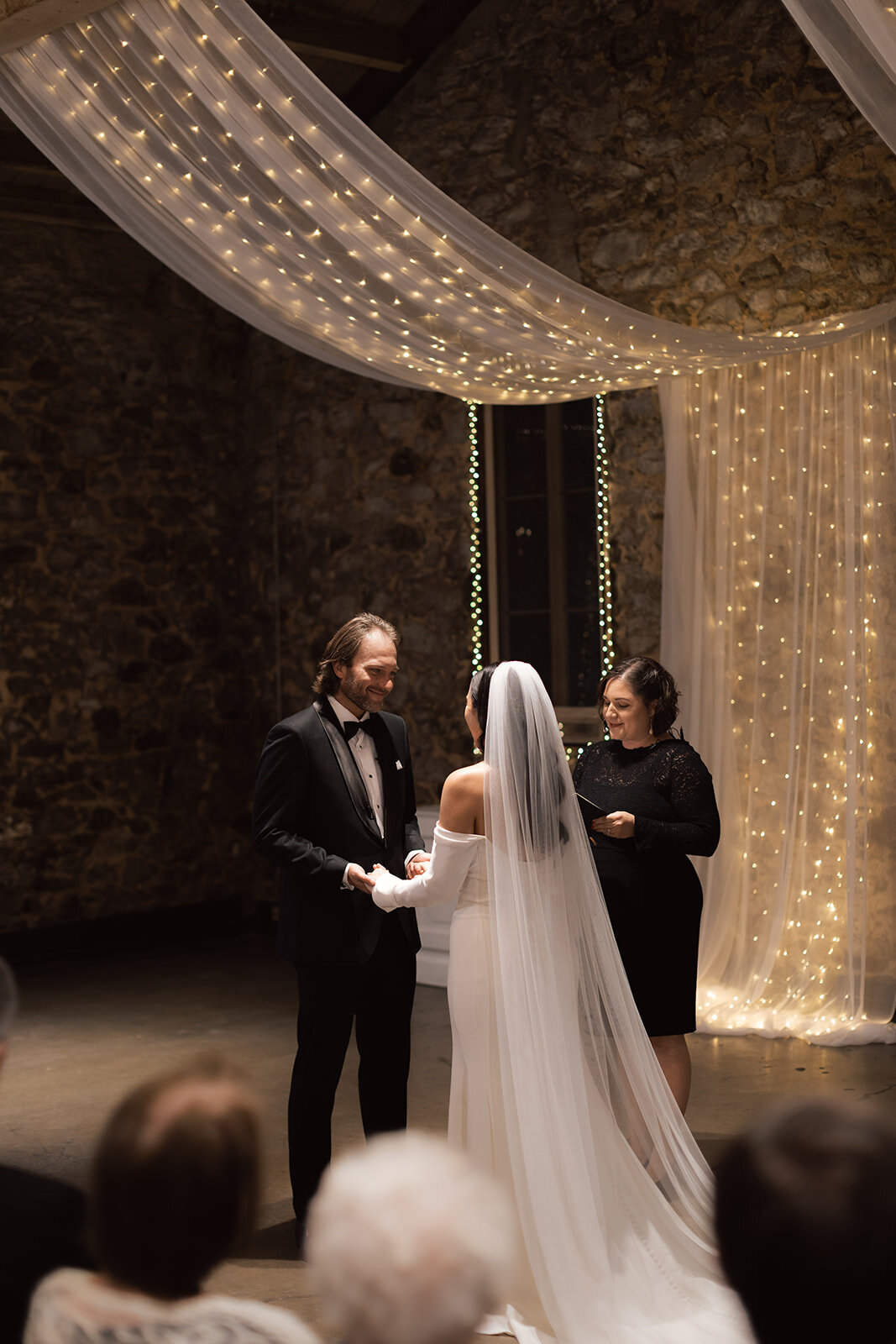 A bride and groom exchange heartfelt vows under twinkling string lights in an intimate wedding ceremony. A romantic and artistic wedding moment captured with warmth.