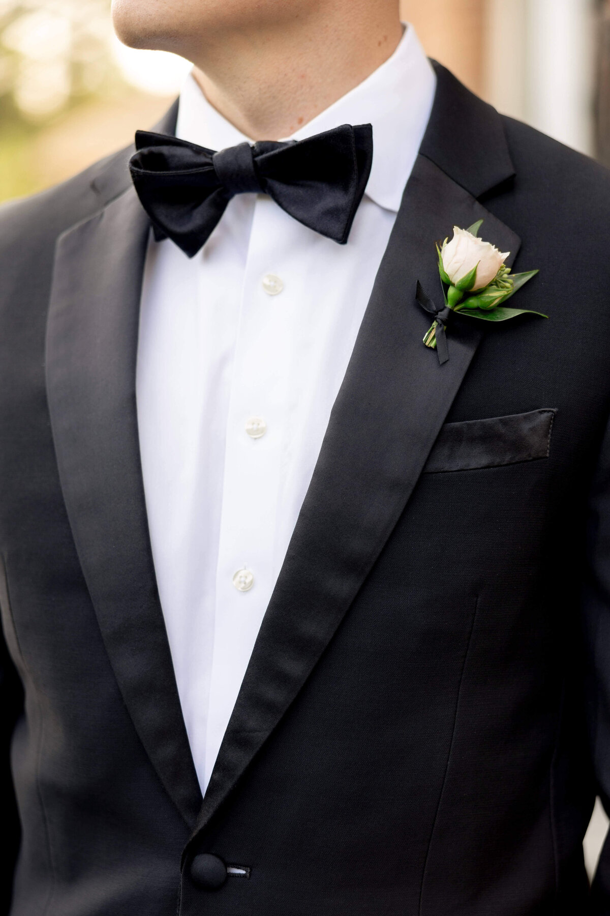 A man in a black tuxedo with a black bow tie and a white dress shirt. A small boutonniere with a pink rose and greenery is pinned to the lapel. The background is softly blurred.