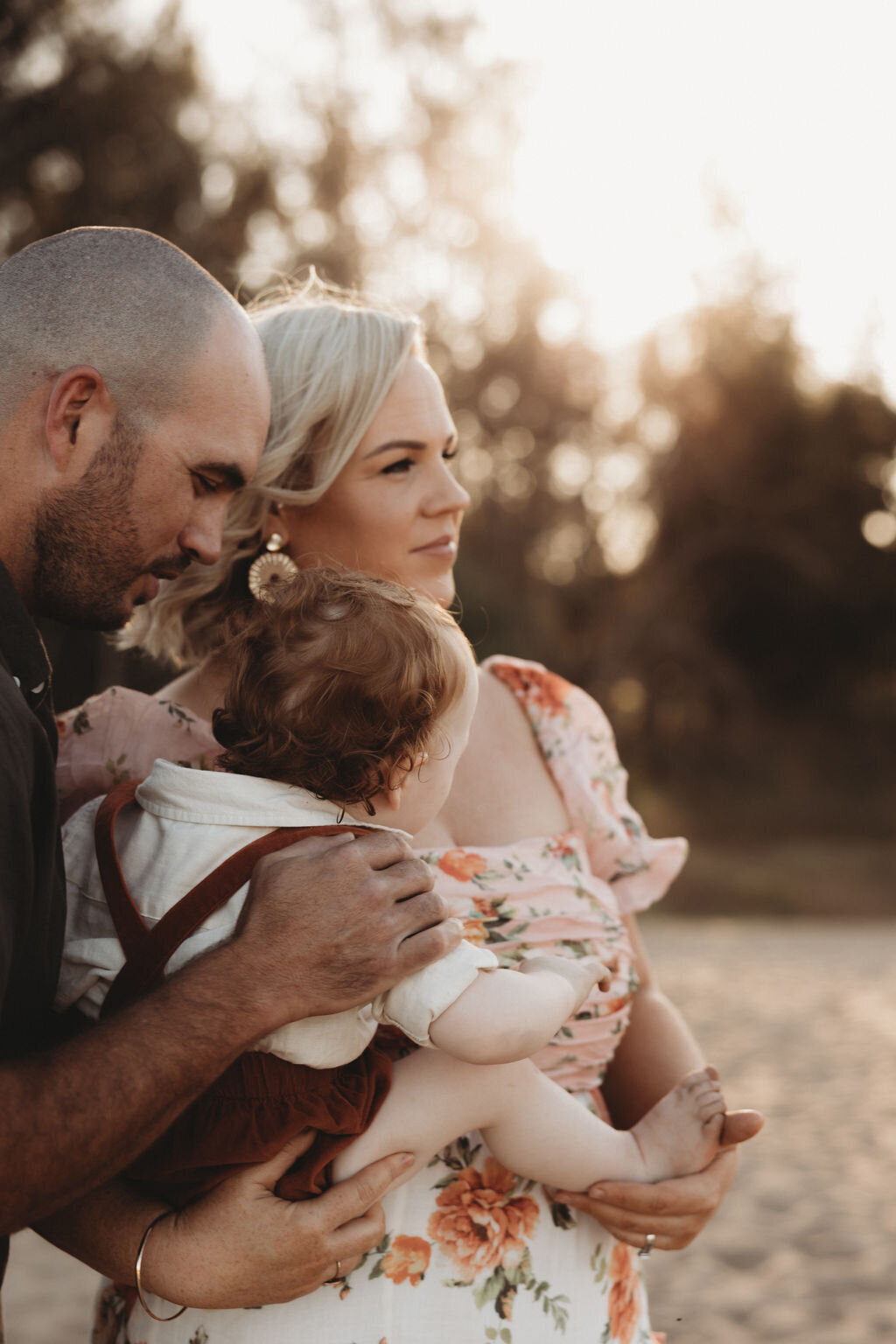 A couple stands together outdoors, holding a baby. The man is on the left side, and the woman is in the middle, with the baby in her arms. Captured by a NSW family photographer, they appear to be looking at something in the distance.