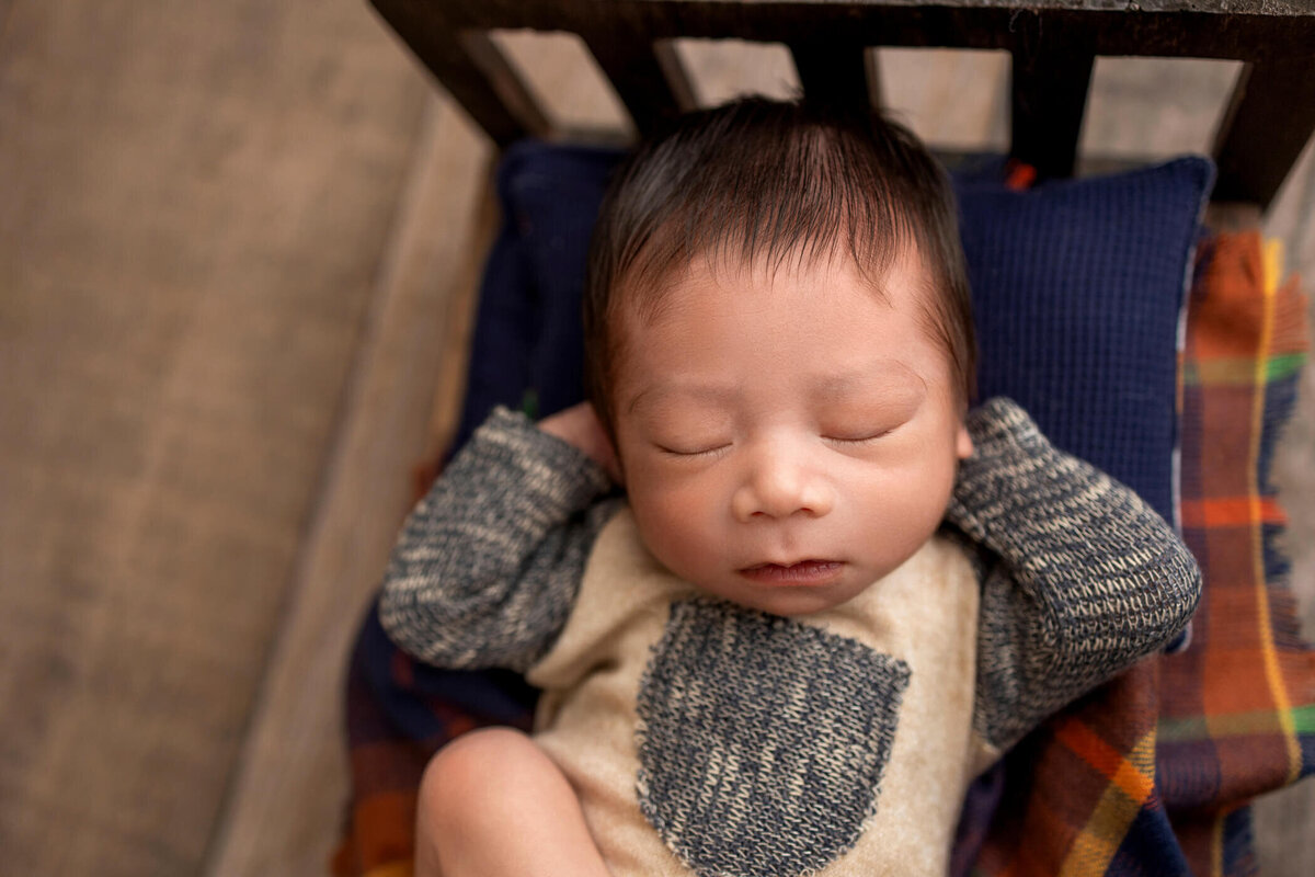 Newborn baby wearing a cozy knit onesie with a bear pattern, sleeping soundly on a plaid blanket and pillow in a wooden crib.