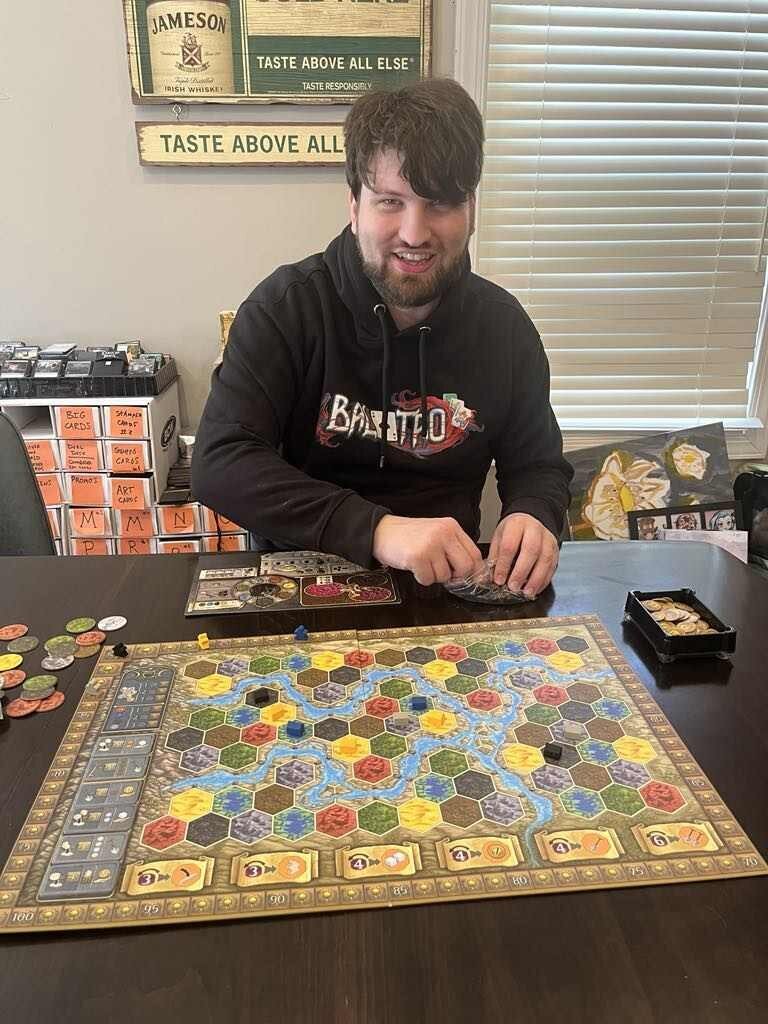 Unpolished dating profile photo of man in hoodie playing a board game with poor lighting.