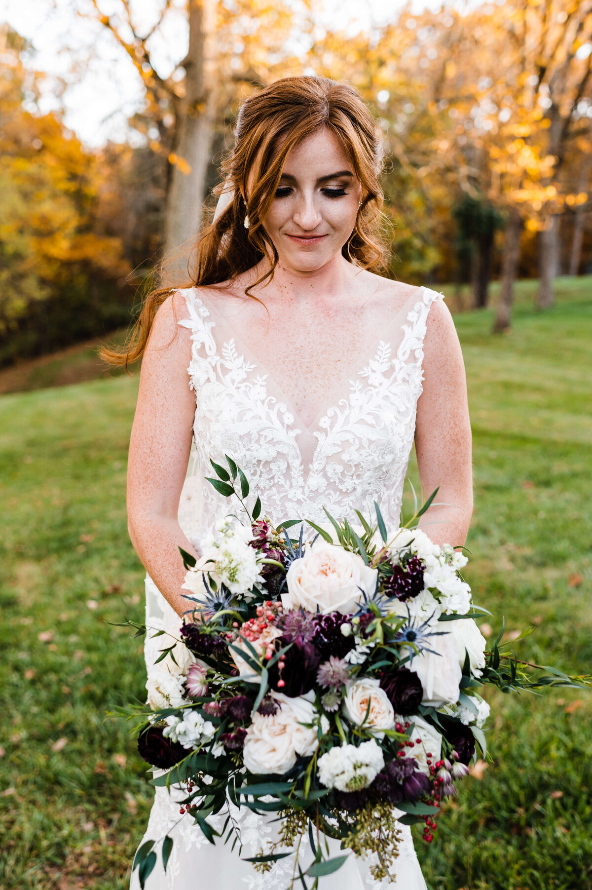 outdoor wedding photo of bride with loose curls framing her face and looking down at her white and purple wedding bouquet that she is holding at her hips while he stands on Charlottesville wedding venues lawn for an autumn wedding