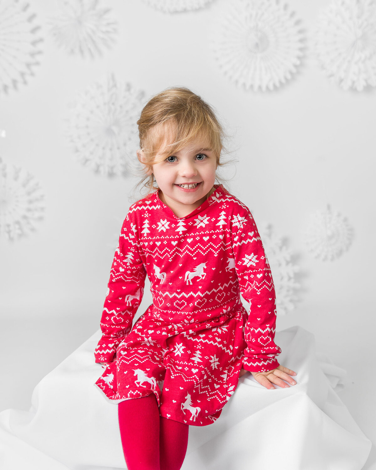 Image of a girl sitting on a large white present on a Christmas snowflake backdrop in the studio. Taken by Norwich portrait photographer Claire Howes. Christmas Mini Sessions Norwich.