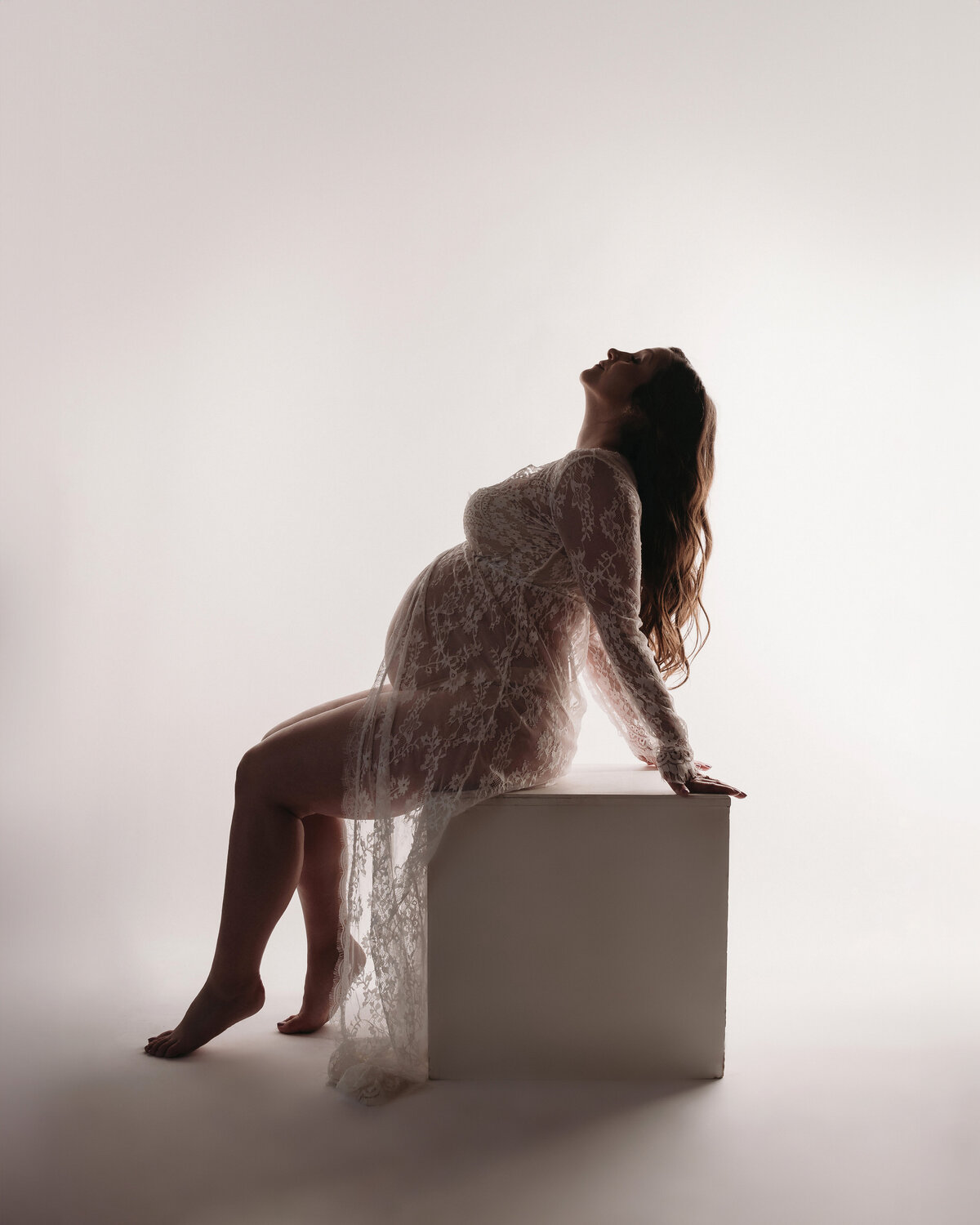 silhouette maternity of pregnant woman wearing white lace gown posing with arched back and hair hanging back on white posing cube in front of white backdrop