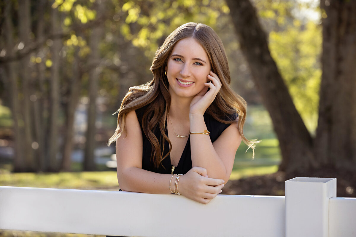 Close up portrait of a smiling high school senior in black dress with white fence and golden trees in Charlotte-area nature setting