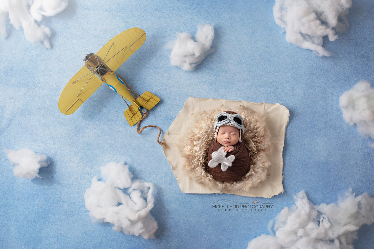 Newborn baby on airplane-themed setup with blue clouds and a yellow plane prop