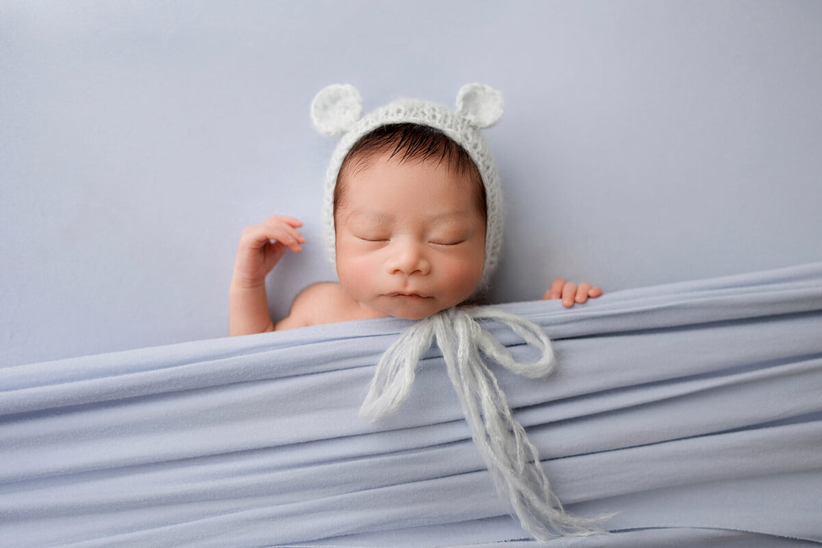 Newborn baby wearing a light blue knit bear hat, peacefully sleeping under a soft blue blanket.