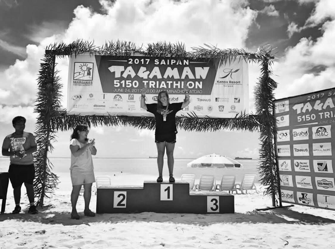 Nicole stands on stage at the 2017 Saipan Tagaman Triathalon winning first place, celebrating with a medal around her neck and her hands in the air.